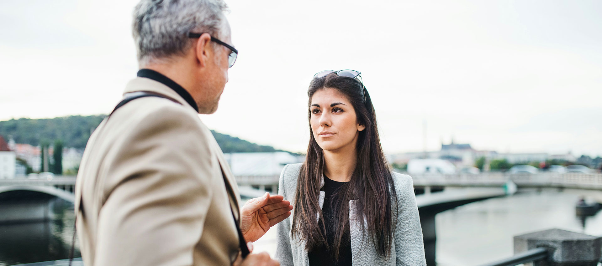 A young woman with long dark hair looks intensely at a middle aged man with grey hair and glasses outside in a busy city.