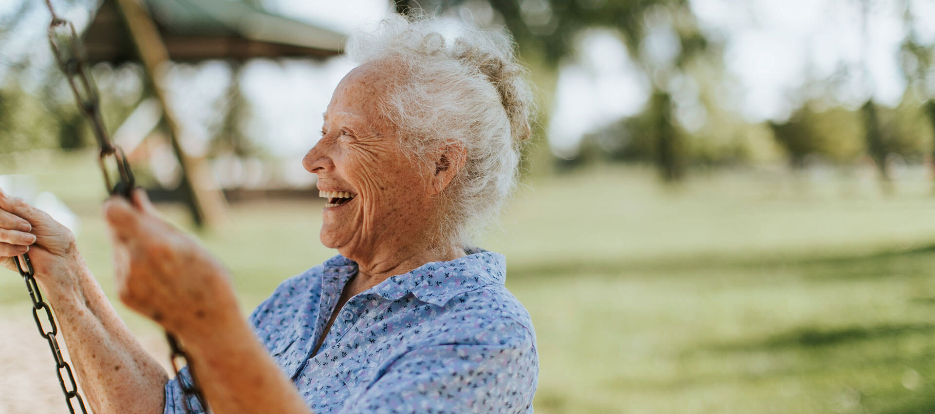 An elderly person in a blue shirt joyfully swings on a swing set in a sunny park, surrounded by green grass and trees.