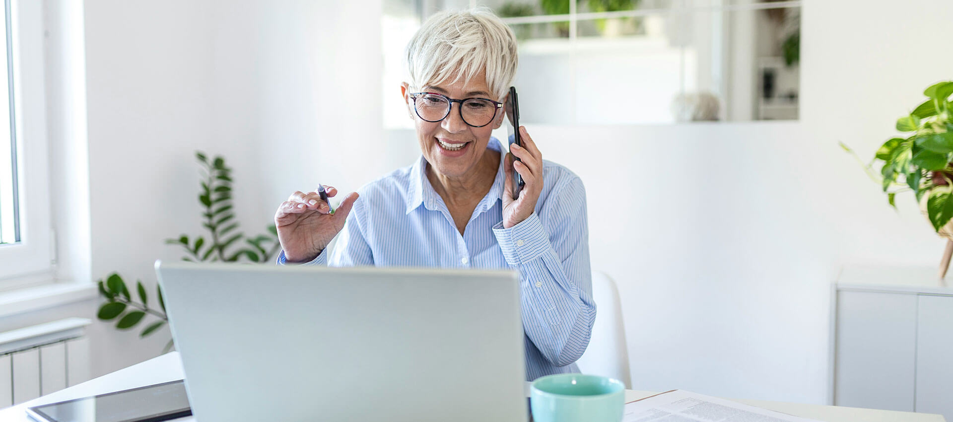 A healthcare professional, wearing glasses and a headset, engages in a video consultation on a laptop. The setting is a bright office with plants in the background.