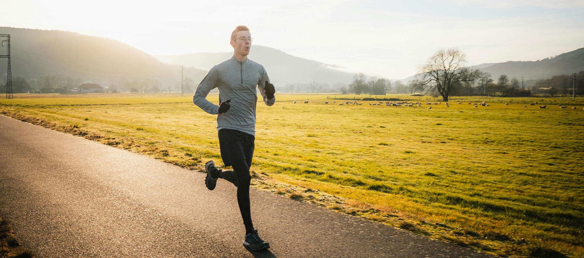 A man in a grey long sleeved running top, black shorts and black skins runs on a paved road in the middle of the country side.