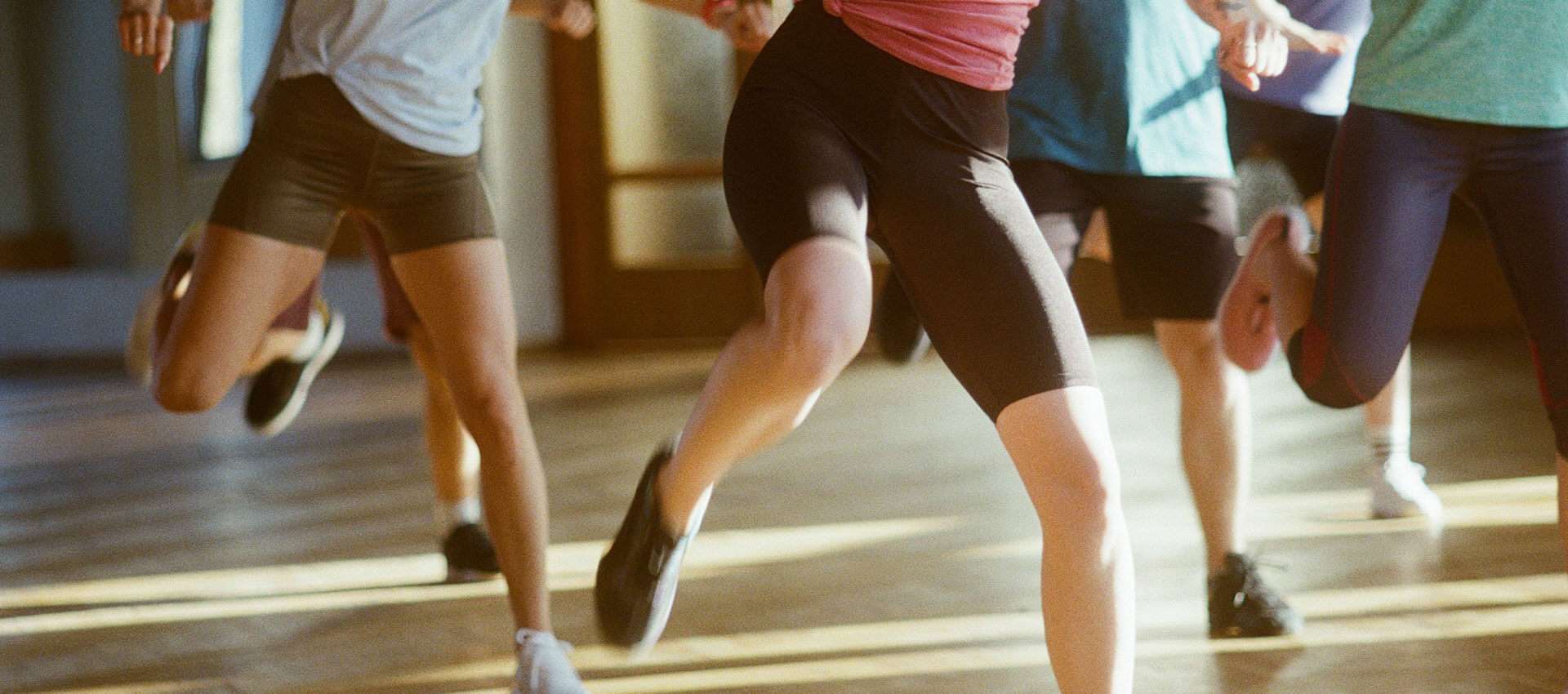 People exercise in a gym, wearing athletic clothing and shoes, moving energetically across a wooden floor. Sunlight streams in through large windows in the background.