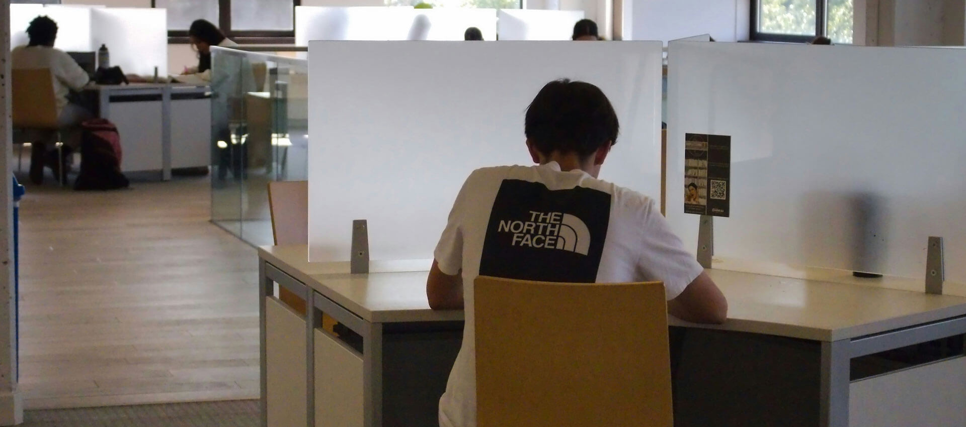 A person in a "The North Face" shirt sits at a partitioned workspace in an open office environment, with others working in the background.