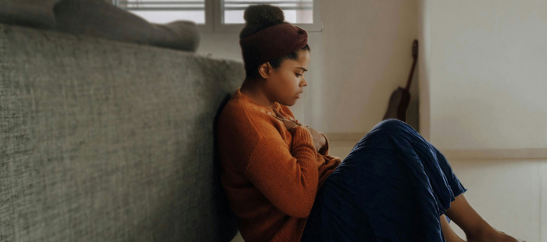 A person sits on the floor, leaning against a sofa with their knees drawn up, in a softly lit room with a guitar and window shutters in the background.
