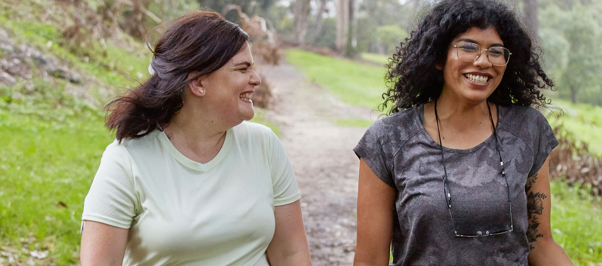 Two people are walking and smiling on a gravel path through a green park. One wears a light shirt, and the other wears a dark patterned shirt and glasses with a lanyard.