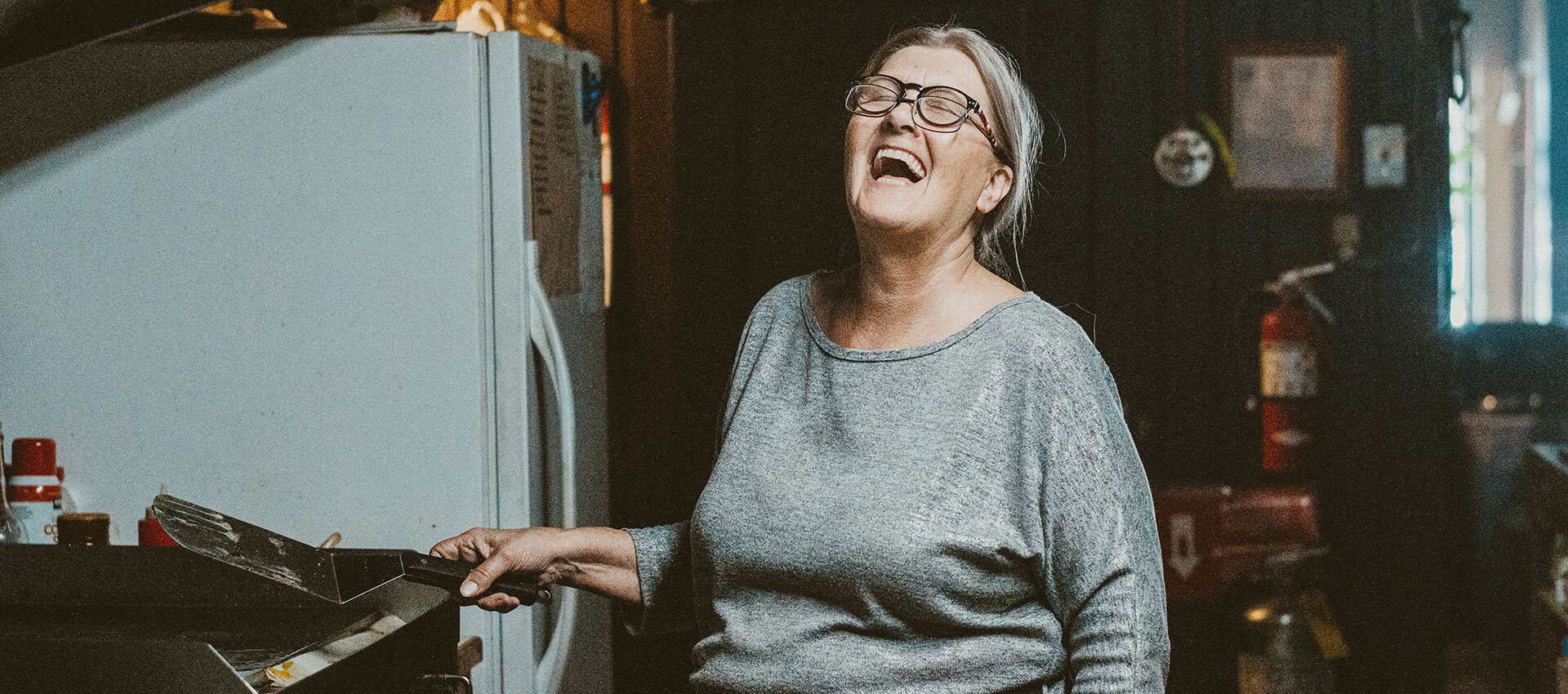 A woman wearing a grey top and glasses laughs with her eyes closed as she stands at a cooker holding a utensil in her hands.
