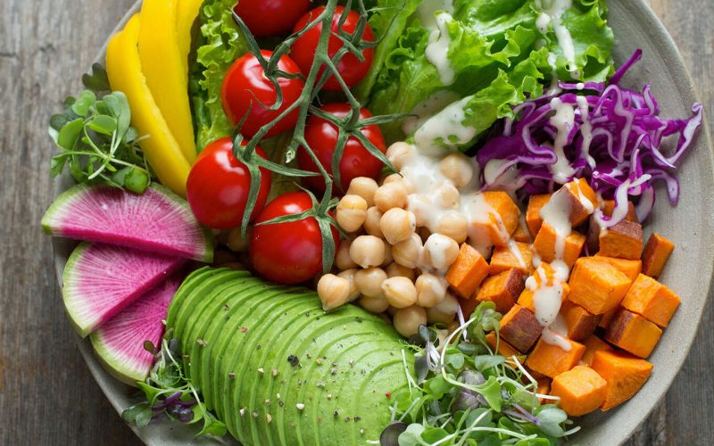 A bowl contains sliced avocado, radishes, cherry tomatoes, yellow bell pepper, chickpeas, cubed sweet potatoes, and lettuce. Drizzled with dressing, it rests on a rustic wooden surface.
