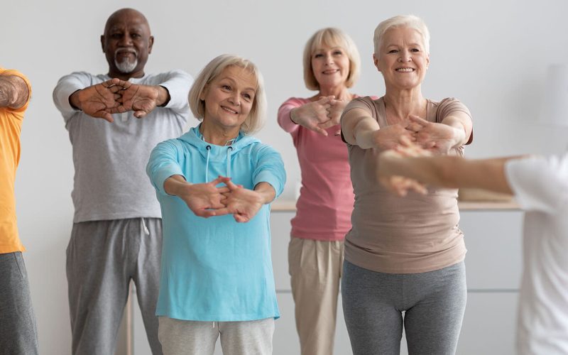 Elderly individuals perform stretching exercises, standing with arms extended forward, in a bright room.