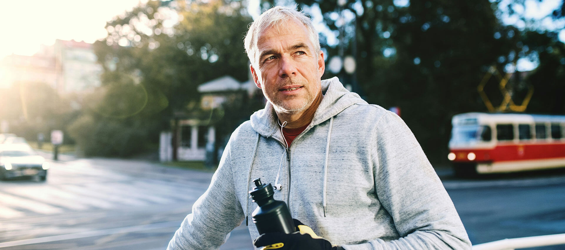 A man, in a gray hoodie holding a water bottle, stands by a street during a sunny day. A vintage tram and trees are visible in the background.