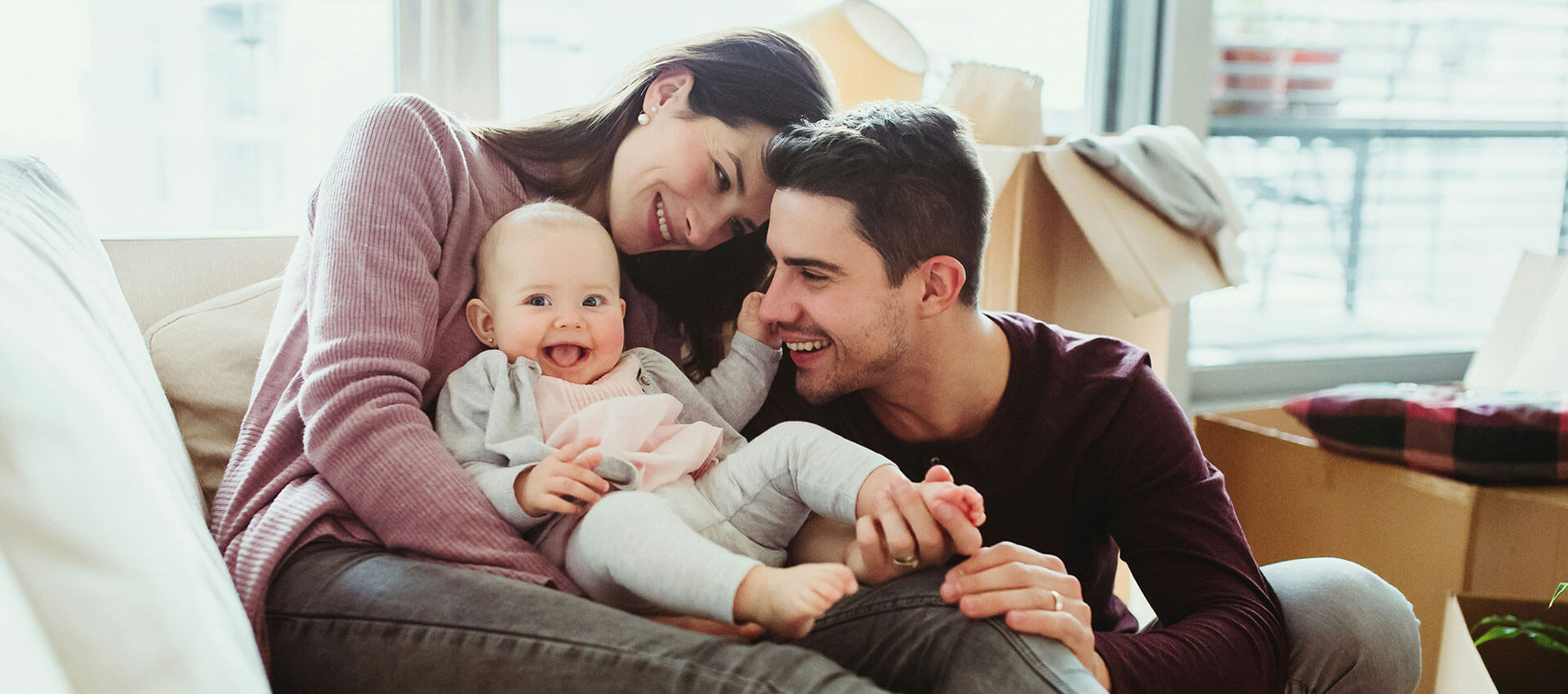 A baby sits on a couch, held affectionately by smiling parents, surrounded by cardboard boxes, suggesting a recent move or unpacking in a bright, modern room.