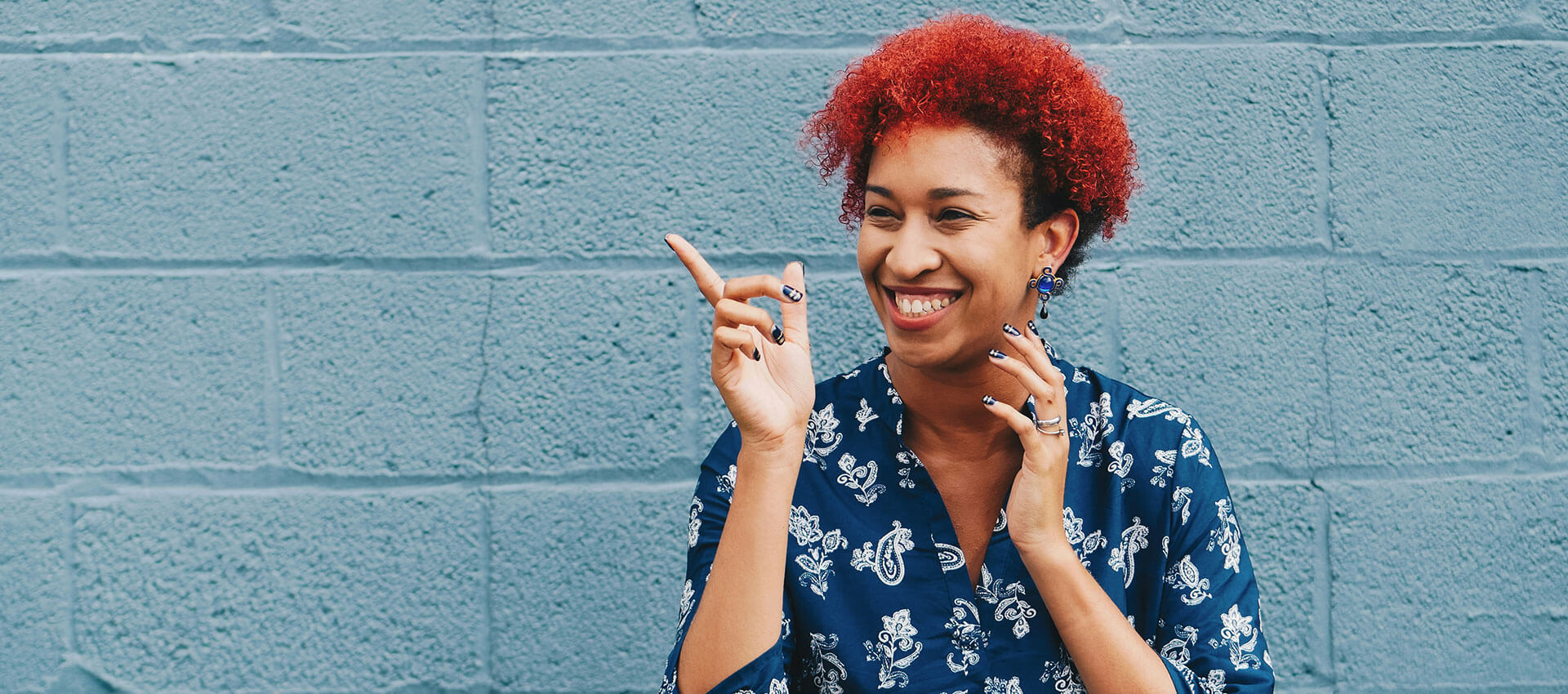 A person with bright red hair smiling and pointing upward, set against a blue brick wall background, wearing a patterned shirt.