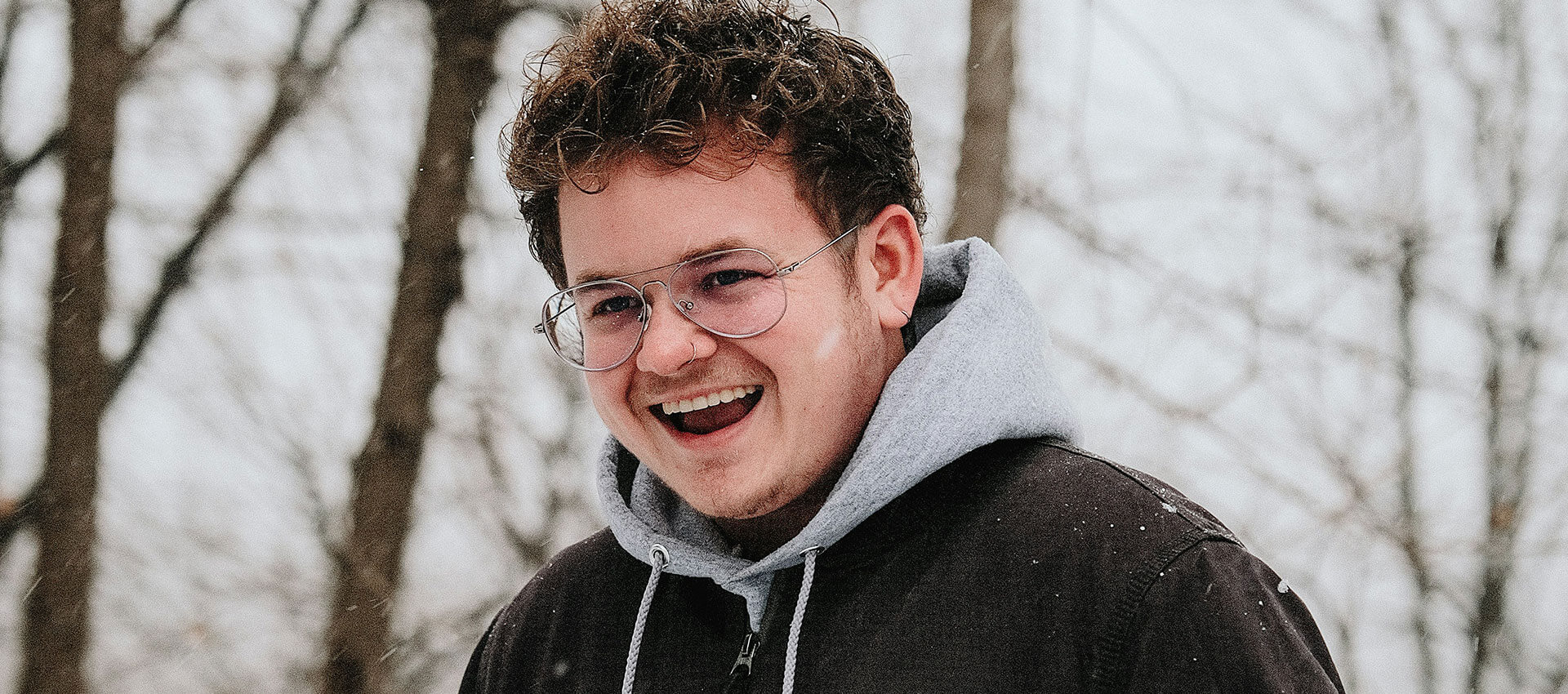 A young man with short curly hair and silver glasses, laughs as he walks through the snow wearing a grey hoody and a black coat.
