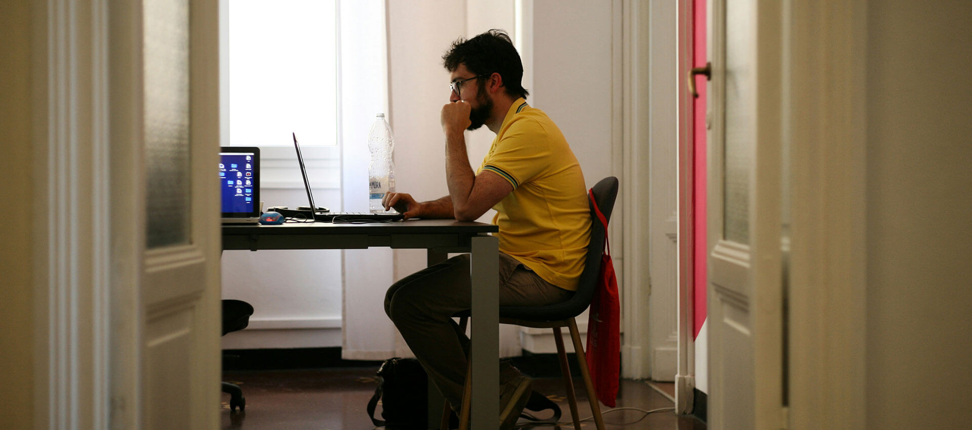 A person in a yellow shirt sits at a desk, using a laptop, in a well-lit room with white walls and a visible bottle of water.
