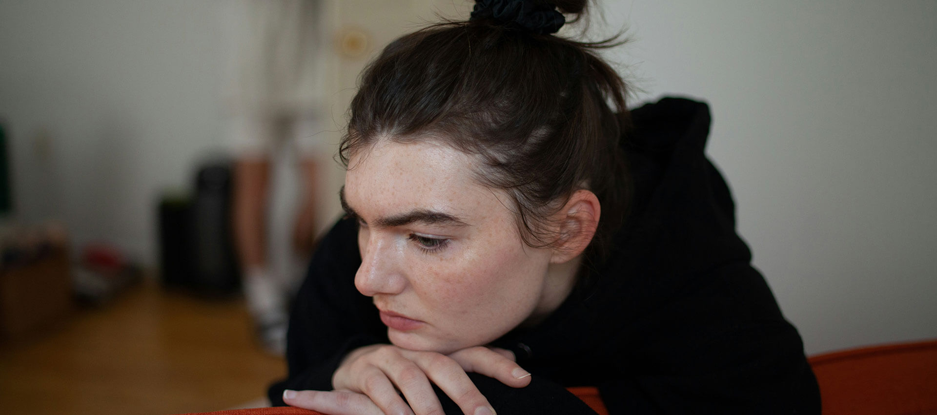 A close up of a woman with her hair ties up in a bun on top of her head, wears a black hoody and looks glum sitting with her chin resting on her hands at a wooden table.