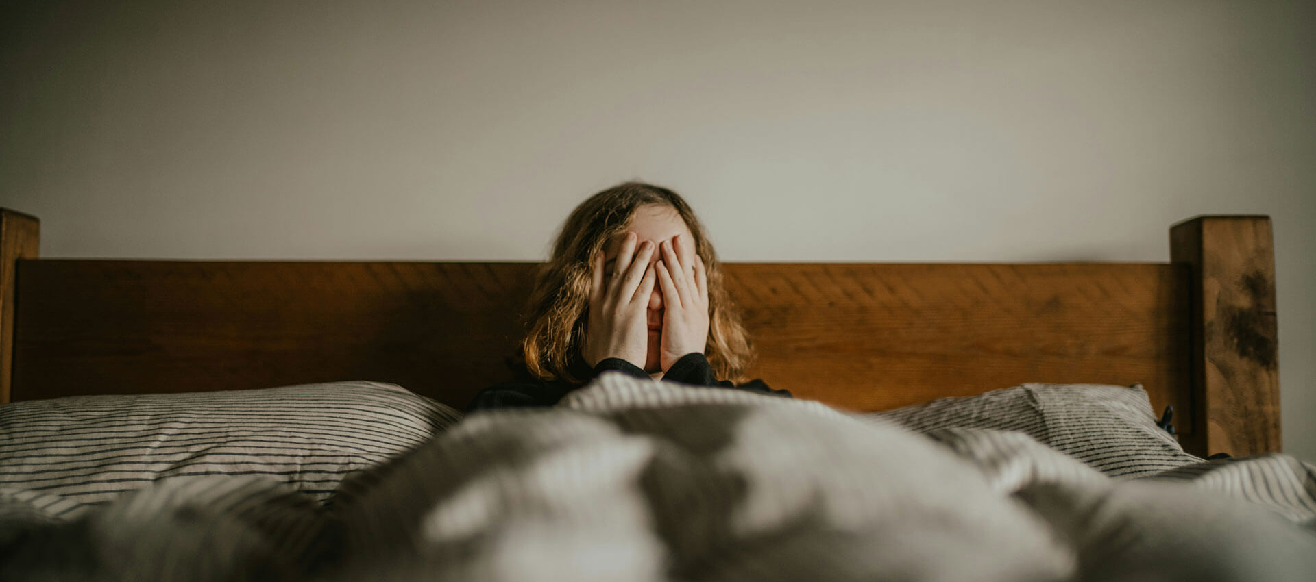 A person with short blonde curly hair sits up in a wooden bed with hands covering their face.