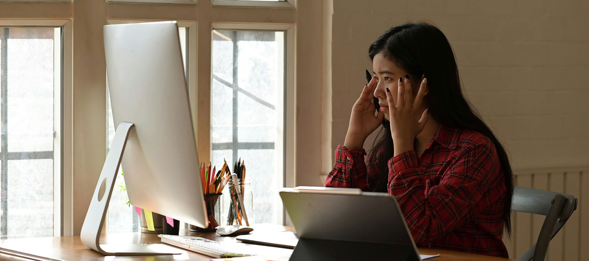 A woman with long dark hair wearing a red and black plaid shirt sits at a large silver computer screen with her head in her hands.