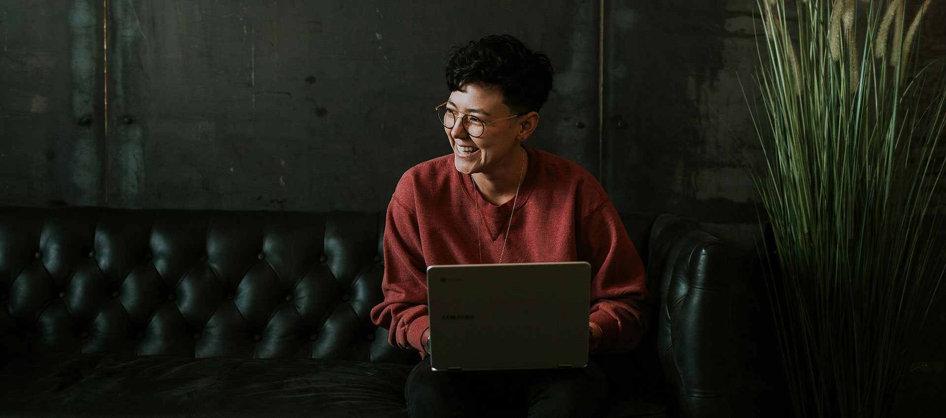 A woman with short dark hair and glasses sits on a black leather sofa with a laptop, laughing at something someone days off camera.