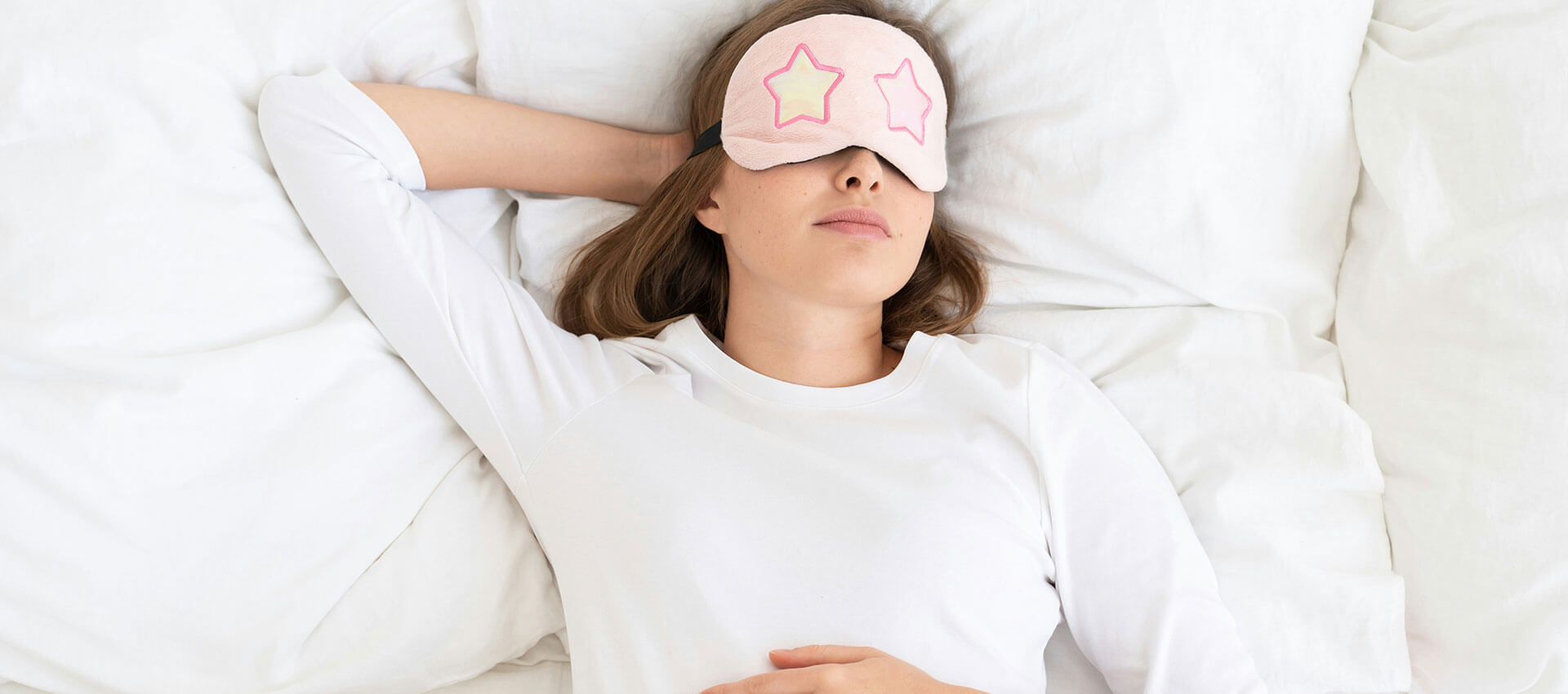 A person is lying in bed, wearing a pink sleep mask with a star design, resting on white pillows and sheets.