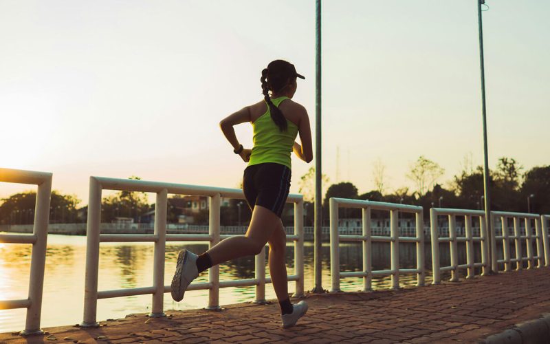 A person runs along a paved path beside a river at sunrise, surrounded by railing and streetlights.