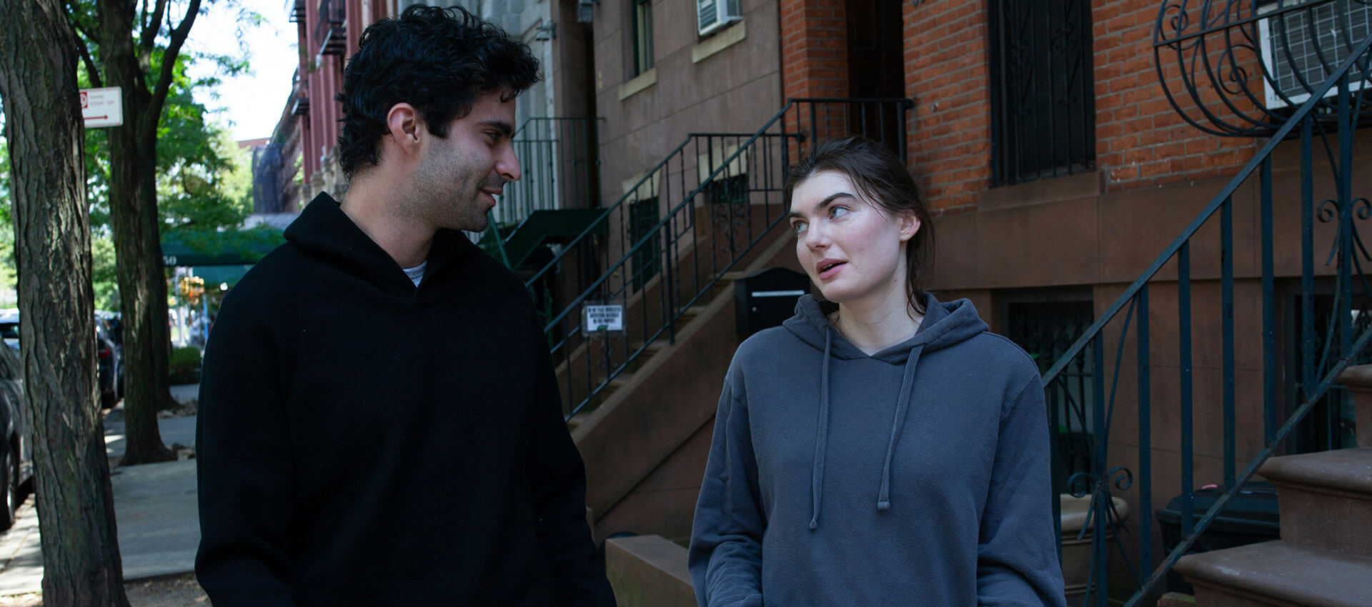 A girl wearing a dark grey hoody talks to a man wearing a black hoody as they walk down a street lined with red brick houses.