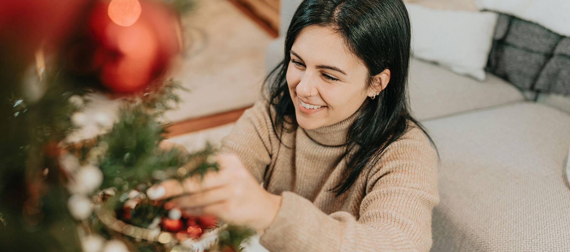 A woman with dark shoulder length hair, sits on the floor in her living room while decorating her Christmas tree with red baubles.