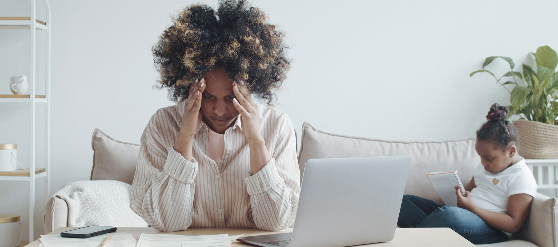 A woman with shirt curly hair sits at a dining room table with her head in hands looking at her laptop and paperwork, whilst a little girl sits on the sofa in the background writing in a notepad.