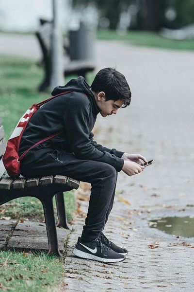 A young person in a black outfit sits on a park bench, looking at a smartphone. A red backpack is slung over their back, and autumn leaves are scattered on the pathway.