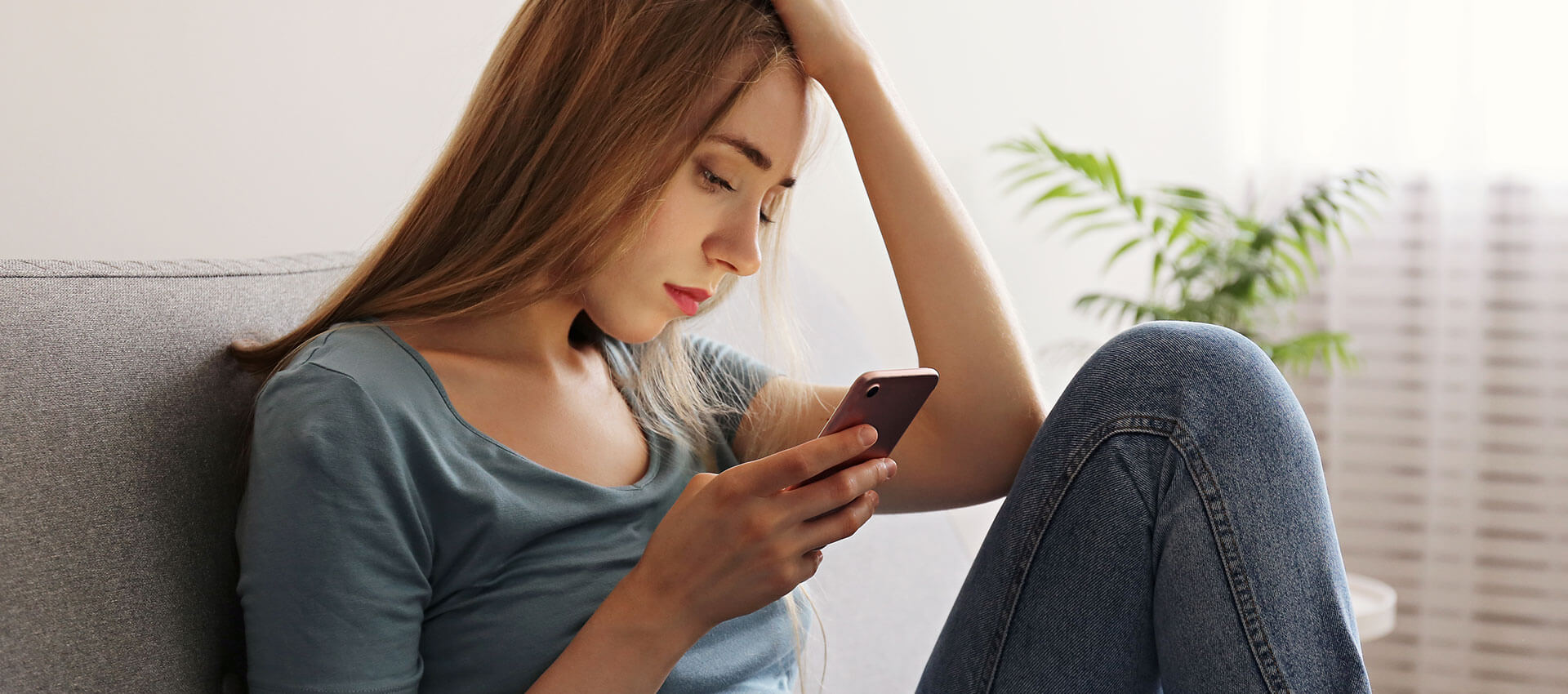 A person seated on a couch looks at a smartphone while holding their head with one hand, in a well-lit room with soft furnishings.