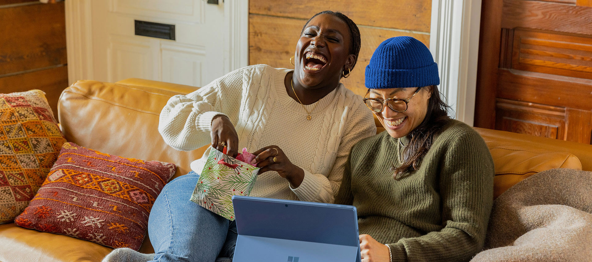 Two women sit on a brown leather couch laughing while on a tablet device.