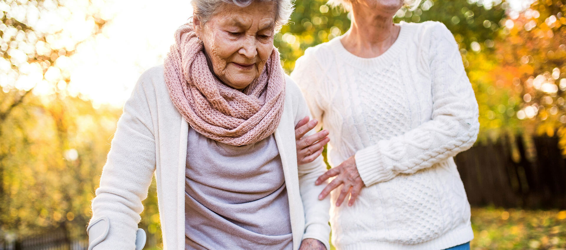 Elderly woman walking outside in a park during Autumn with a walking stick in her right hand and a woman holding her left arm.