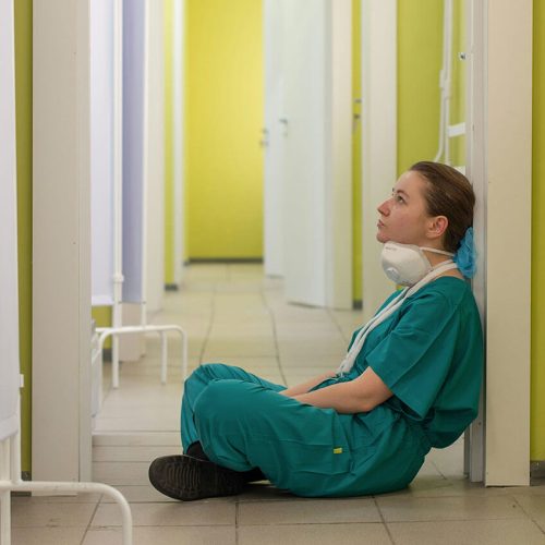 A nurse in green scrubs wearing a medical mask sits crossed legged on the floor of a changing room staring at the wall.