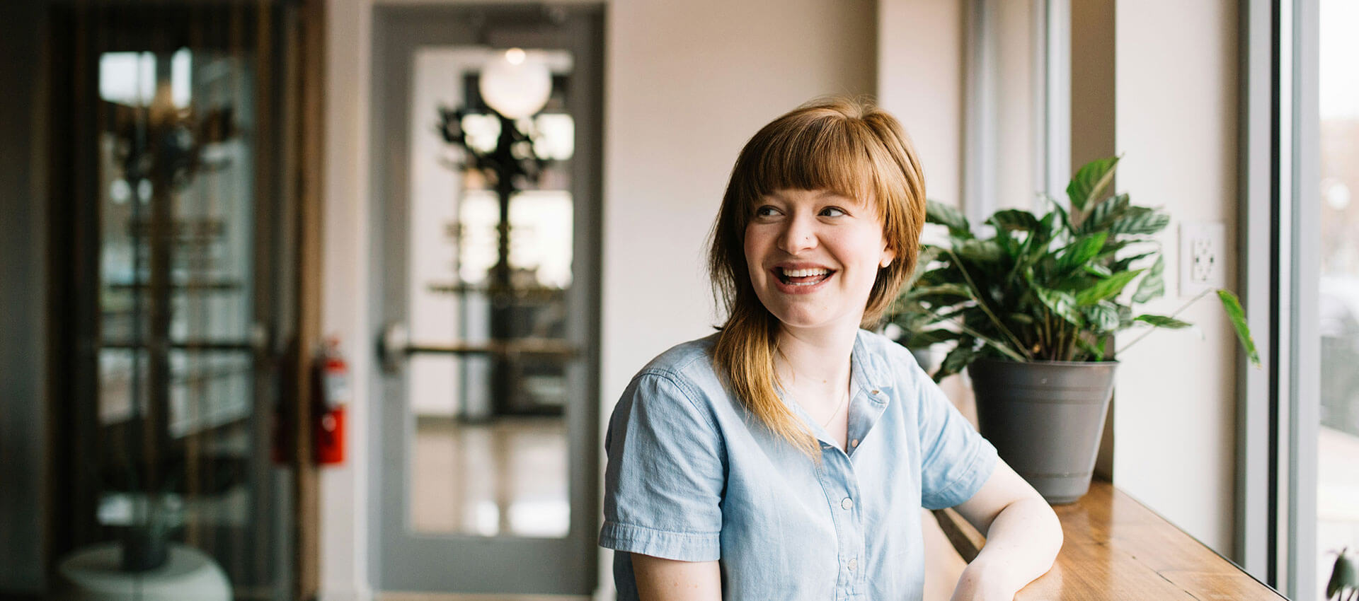 A person is sitting and smiling by a window with natural light, next to a potted plant on a wooden surface in a modern indoor setting.