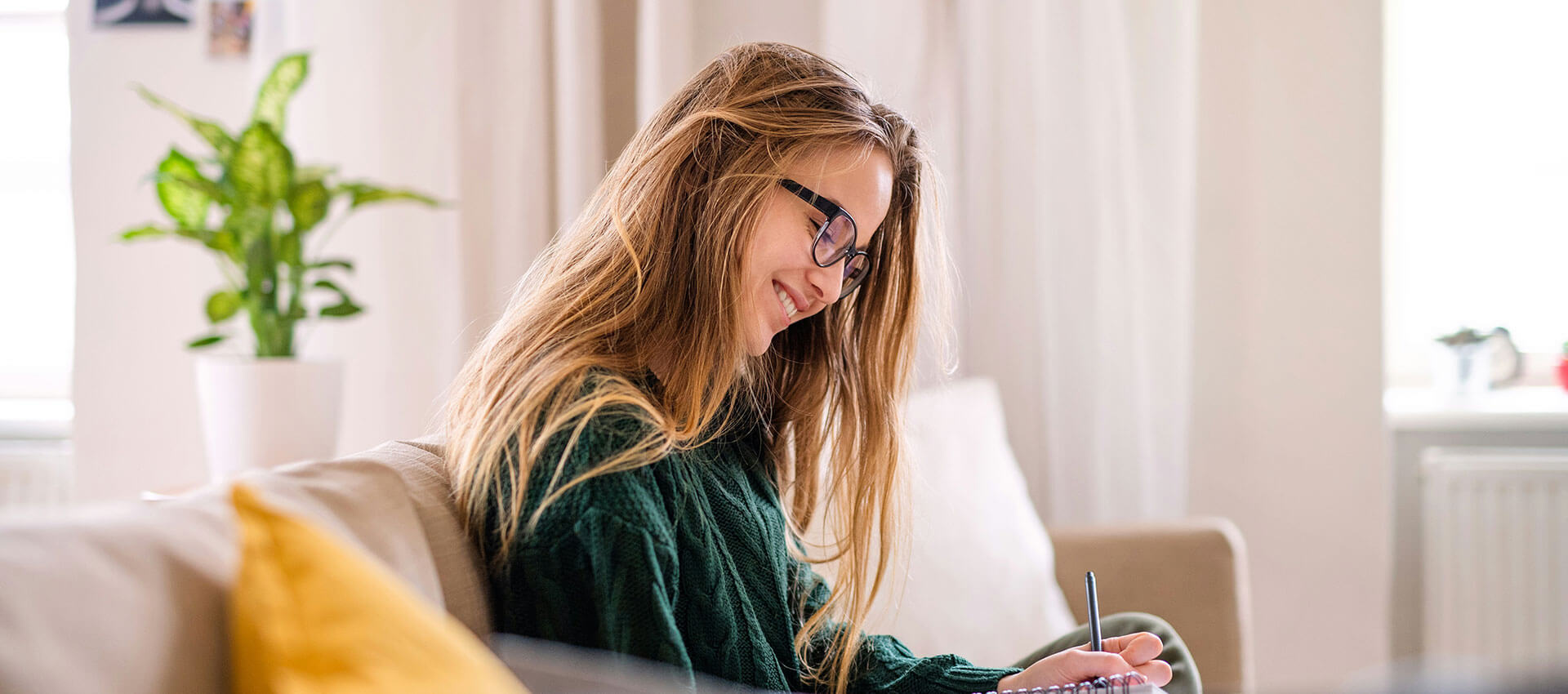 A woman with long hair and glasses sits on a couch, writing in a notebook. She is in a bright, cozy room with a potted plant in the background.