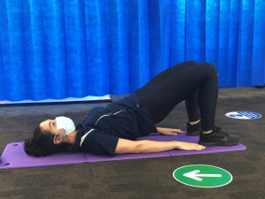 A physiotherapist performs a bridge exercise on a purple mat. They are wearing a face mask. The background features a blue curtain, with directional floor signs visible.