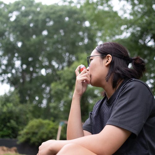 A person in a dark t-shirt sits outdoors, resting their chin on their hand, looking thoughtful. They are surrounded by trees and greenery in a park-like setting.