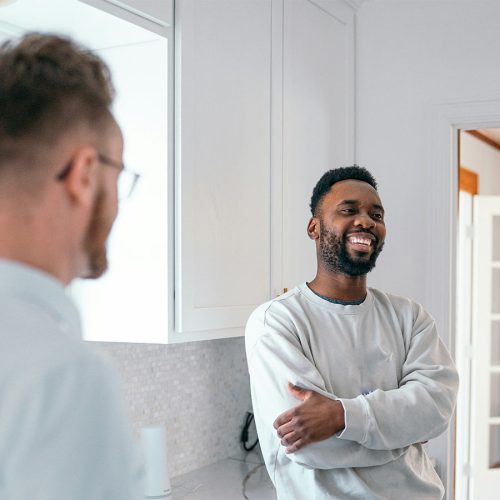 A man smiles with arms crossed while talking to another man in a bright kitchen with white cabinets and tiled backsplash.