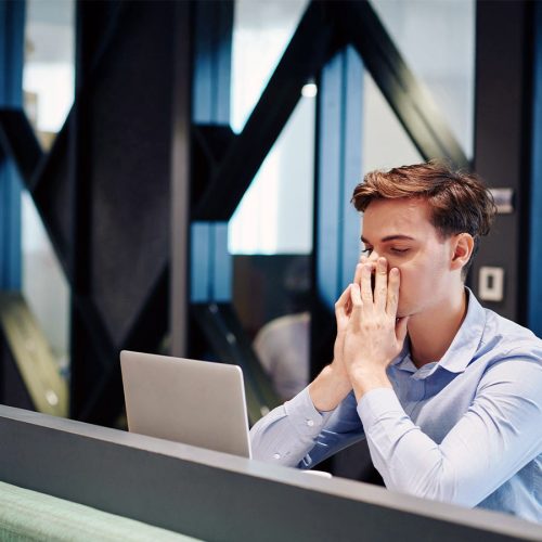 A man sits at a desk with a laptop, covering his mouth with clasped hands. He appears thoughtful or stressed in a modern office environment.