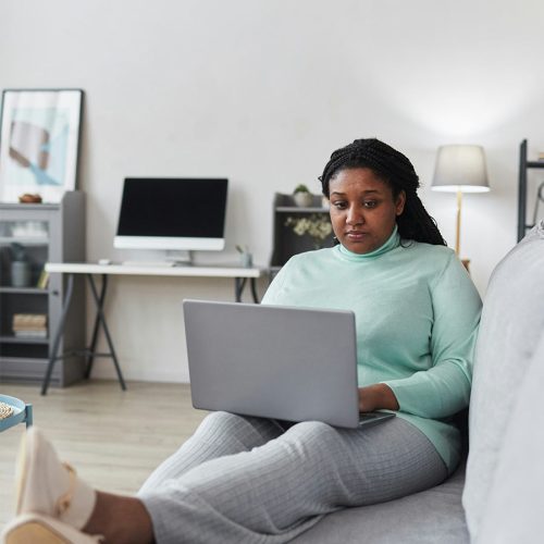 A person in a light blue sweater sits on a couch using a laptop. In the background, a desk with a computer and a lamp is visible, creating a home office setting.