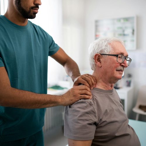 A healthcare professional assists an elderly man with shoulder exercises in a brightly lit room with medical equipment in the background.