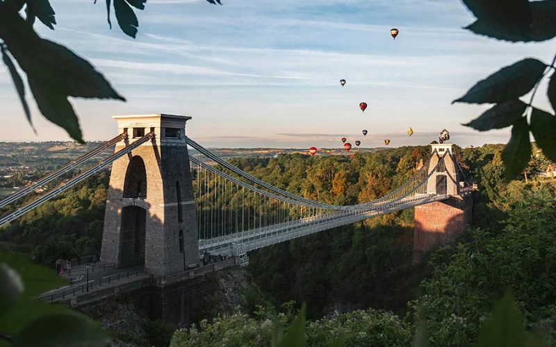A suspension bridge spans across a deep green valley with several colorful hot air balloons floating in the sky. Trees and foliage surround the bridge under a clear blue sky.