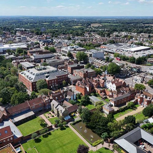 Aerial view of an urban area with a mix of historic and modern buildings surrounded by green spaces and trees, set under a clear blue sky with distant horizon visible.