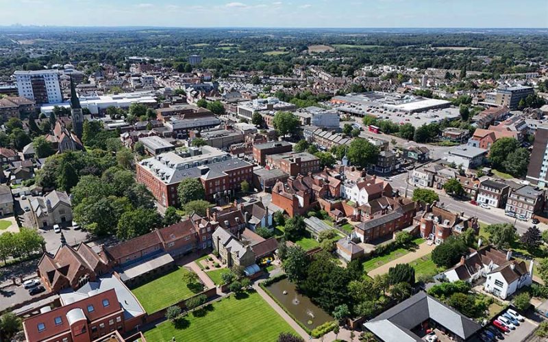 Aerial view of an urban area with a mix of historic and modern buildings surrounded by green spaces and trees, set under a clear blue sky with distant horizon visible.