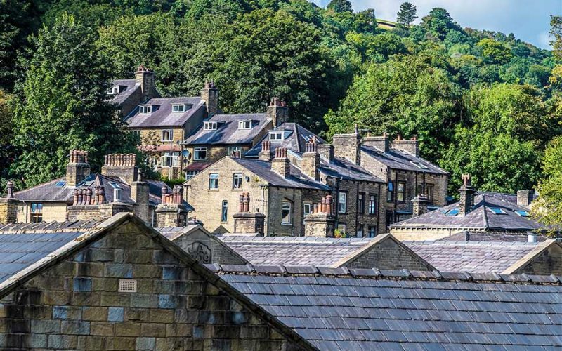 Stone houses with grey slate roofs rest on a hillside, surrounded by lush green trees and a forested background. Sunlight highlights the textures and patterns of the traditional architecture.