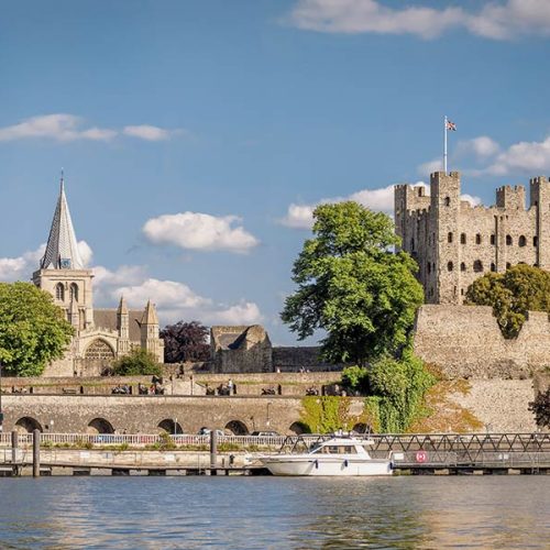 A historic stone castle and church with tall spires stand beside a river. A boat navigates the calm water in the foreground, with trees partially obscuring the structures.