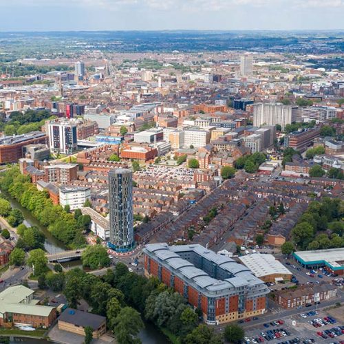 Aerial view of a cityscape with a mix of tall, modern buildings and older structures. Tree-lined streets and a visible river weave through the urban environment under a partly cloudy sky.