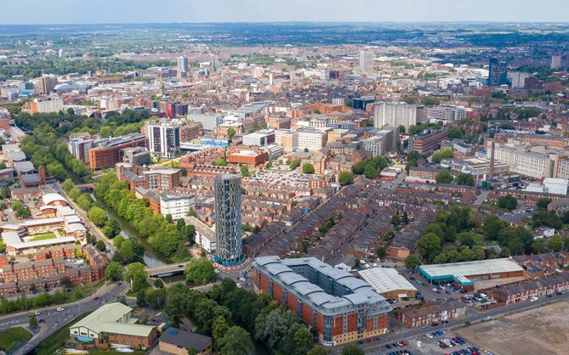 Aerial view of a cityscape with a mix of tall, modern buildings and older structures. Tree-lined streets and a visible river weave through the urban environment under a partly cloudy sky.