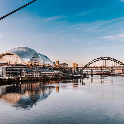 A calm river reflects a modern building with a silver dome and an arch bridge in the background, under a clear blue sky at sunset.
