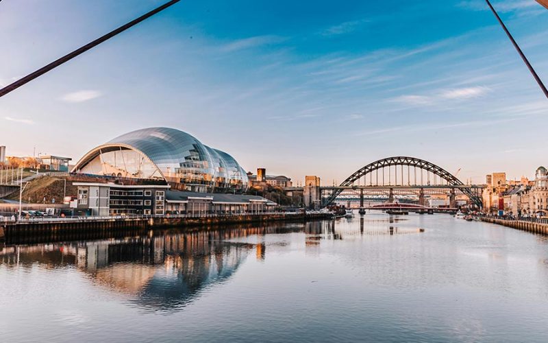 A calm river reflects a modern building with a silver dome and an arch bridge in the background, under a clear blue sky at sunset.