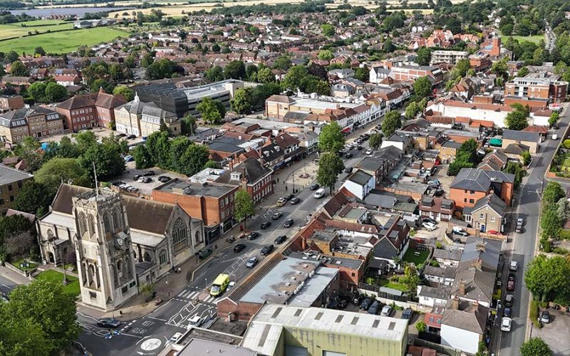Aerial view of a bustling town with scattered cars on streets. Buildings, including a prominent church, are surrounded by trees, with fields visible in the background.