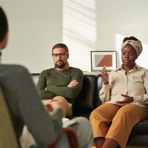 Three people are seated in a room with light-colored walls. One person is speaking while the others listen attentively, suggesting a counseling or discussion session. No text present.