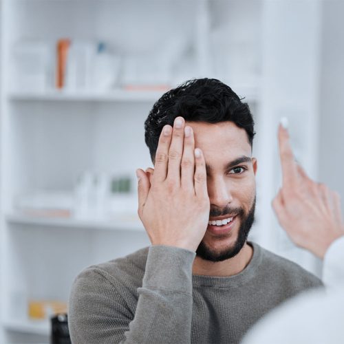 A man covers one eye during an eye examination, smiling slightly as a healthcare professional gestures with a finger. The setting is a bright, clinical space with shelves in the background.
